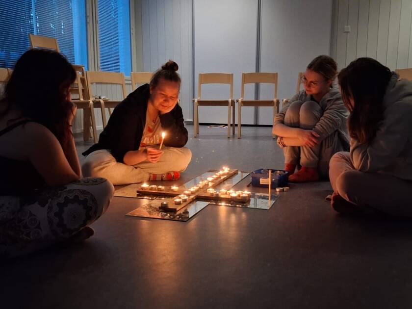 Ohjaaja Mirja ja kolme nuorta kynttilänvalossa, tuikkukynttilät on aseteltu lattialle ristinmuotoon.